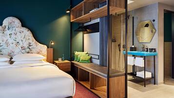 A modern hotel room with a bed, a bench, a shower area, and a vanity with a mirror.