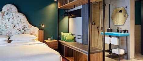 A modern hotel room with a bed, a bench, a shower area, and a vanity with a mirror.