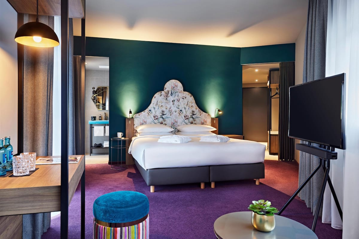 A hotel room with a bed, a TV, a desk with a chair, and a bathroom visible through an open door.