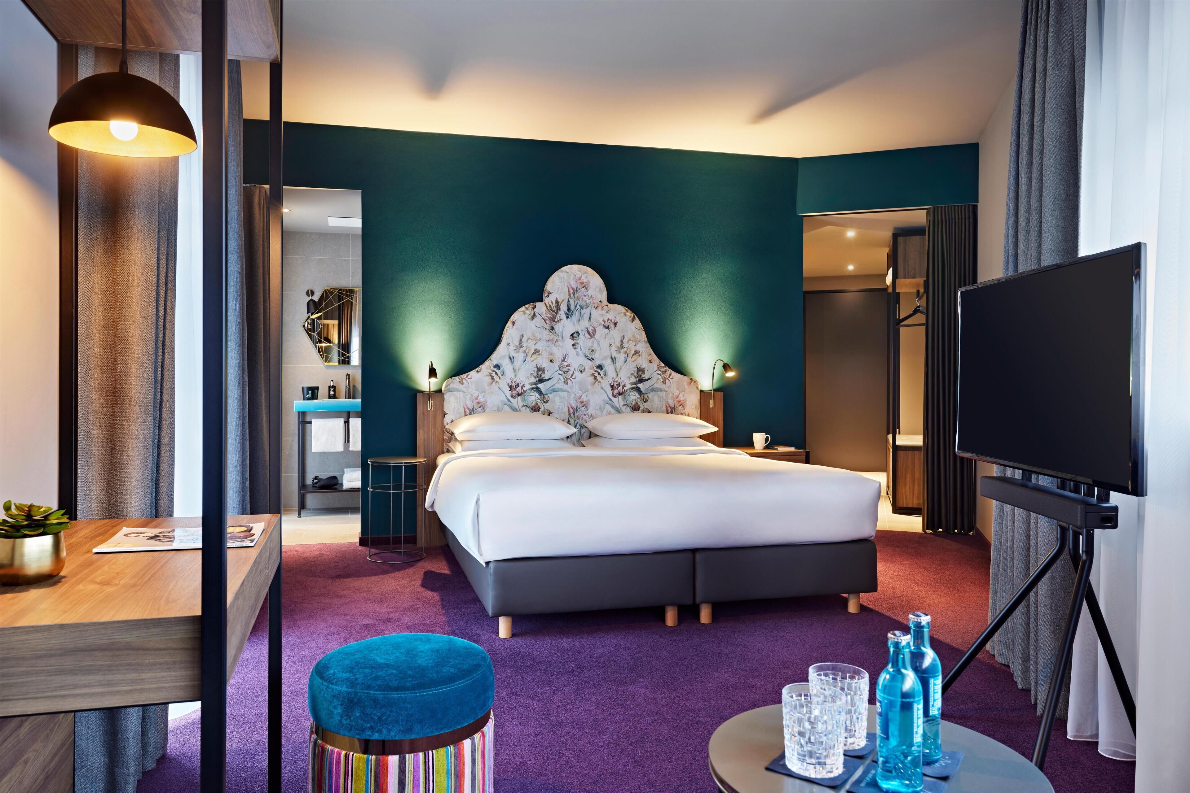 Habitación de hotel con cama, escritorio, televisión y baño visible a través de una puerta abierta.