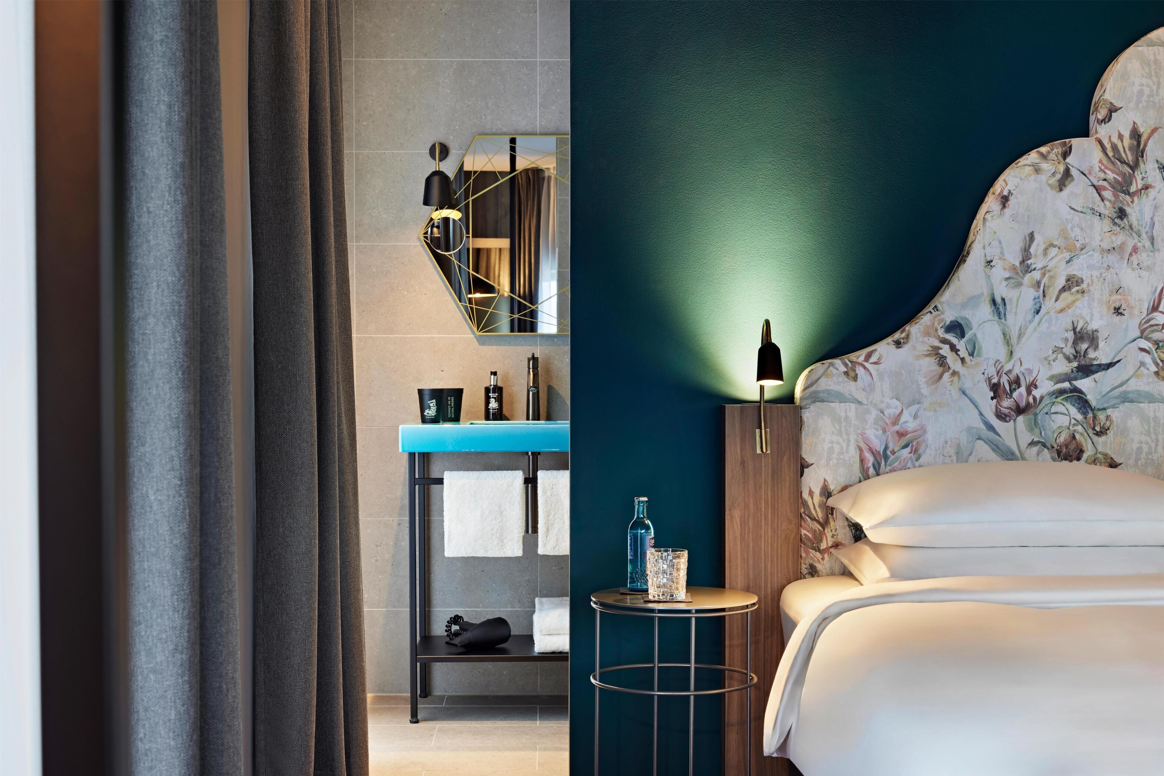 A hotel room with a bed, a small table, a nightstand, a mirror, and a blue wall.