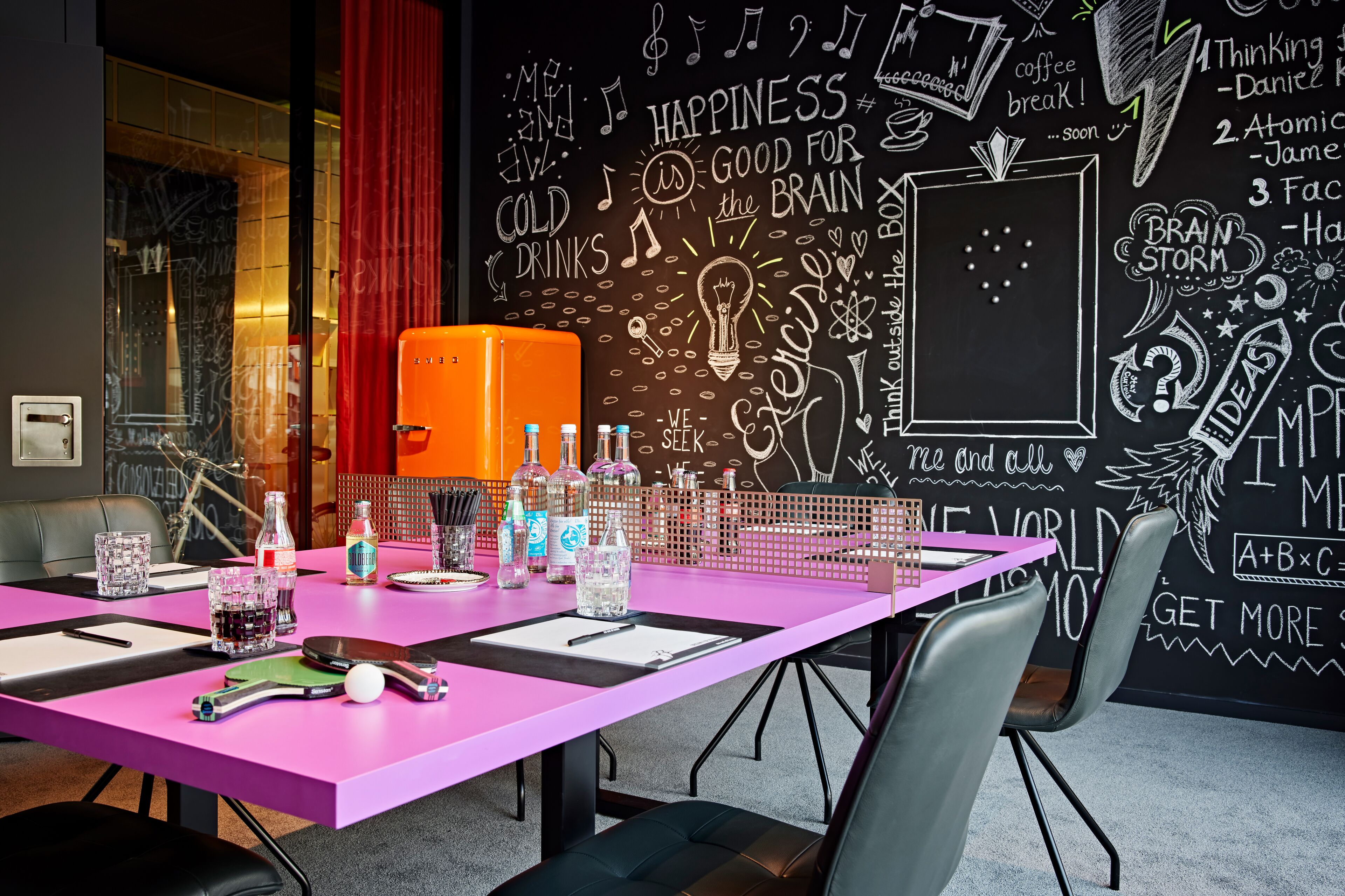 A brightly colored meeting room with a chalkboard wall filled with inspirational doodles and text, a pink table with various items, and a red curtain.