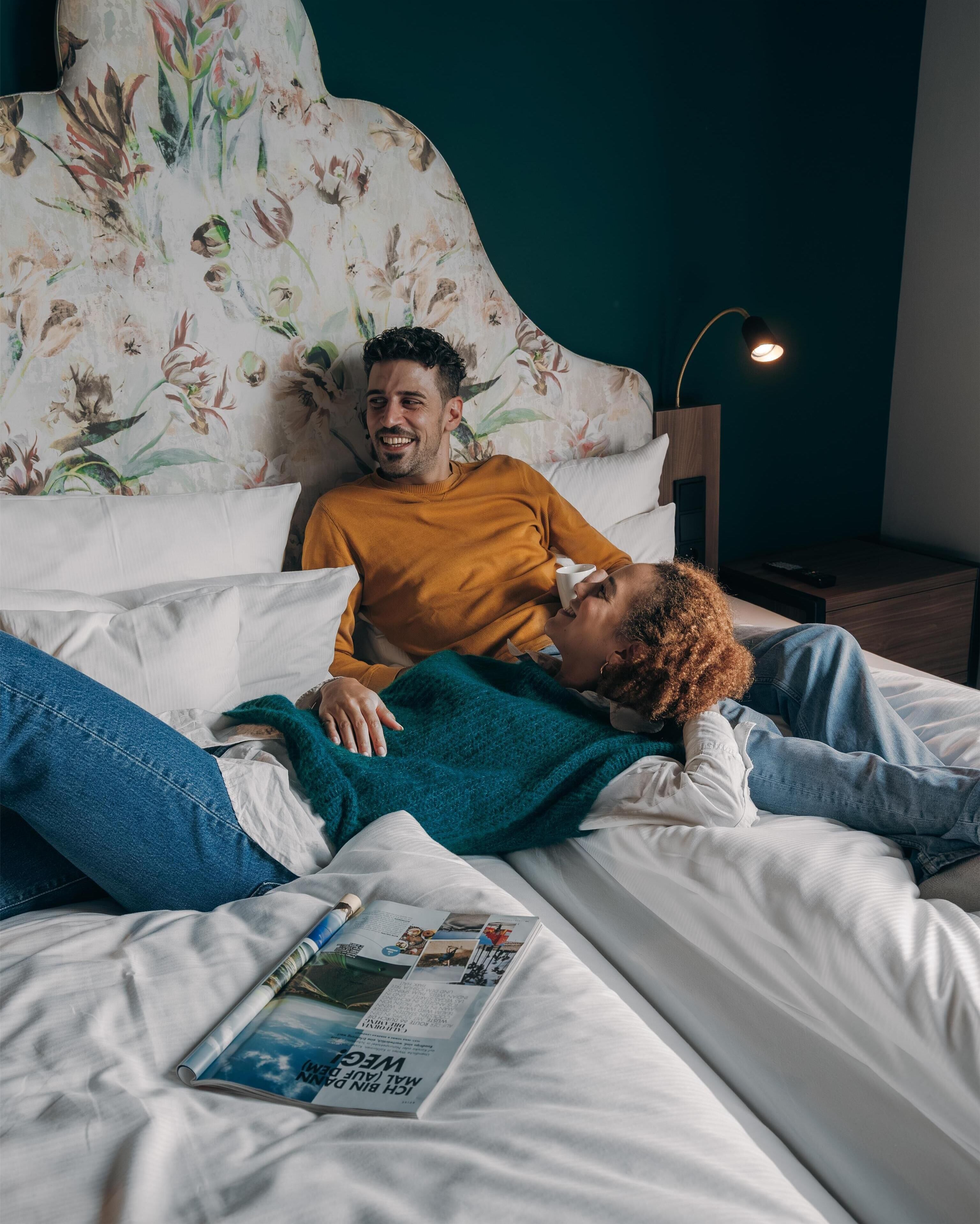 A bedroom with a bed, pillows, a nightstand, a lamp, and a magazine on the bed.