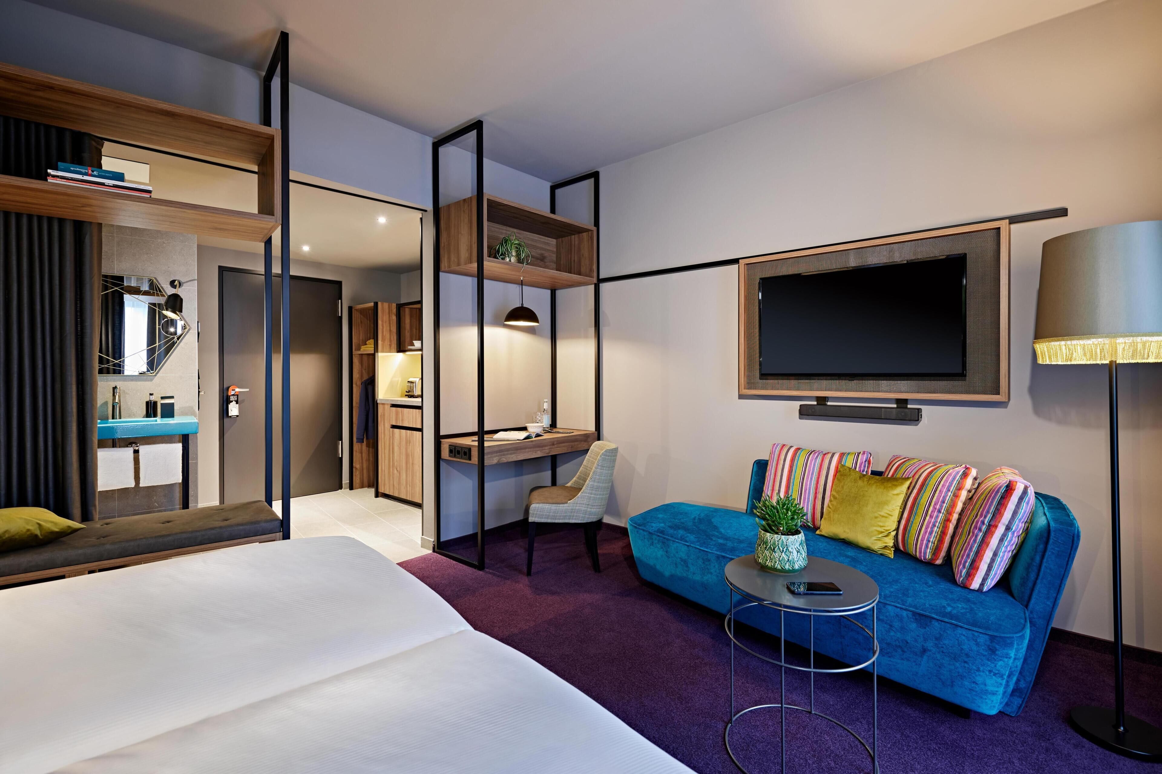 Una habitación de hotel moderna con una cama, un sofá azul, un escritorio, un televisor y un espejo.