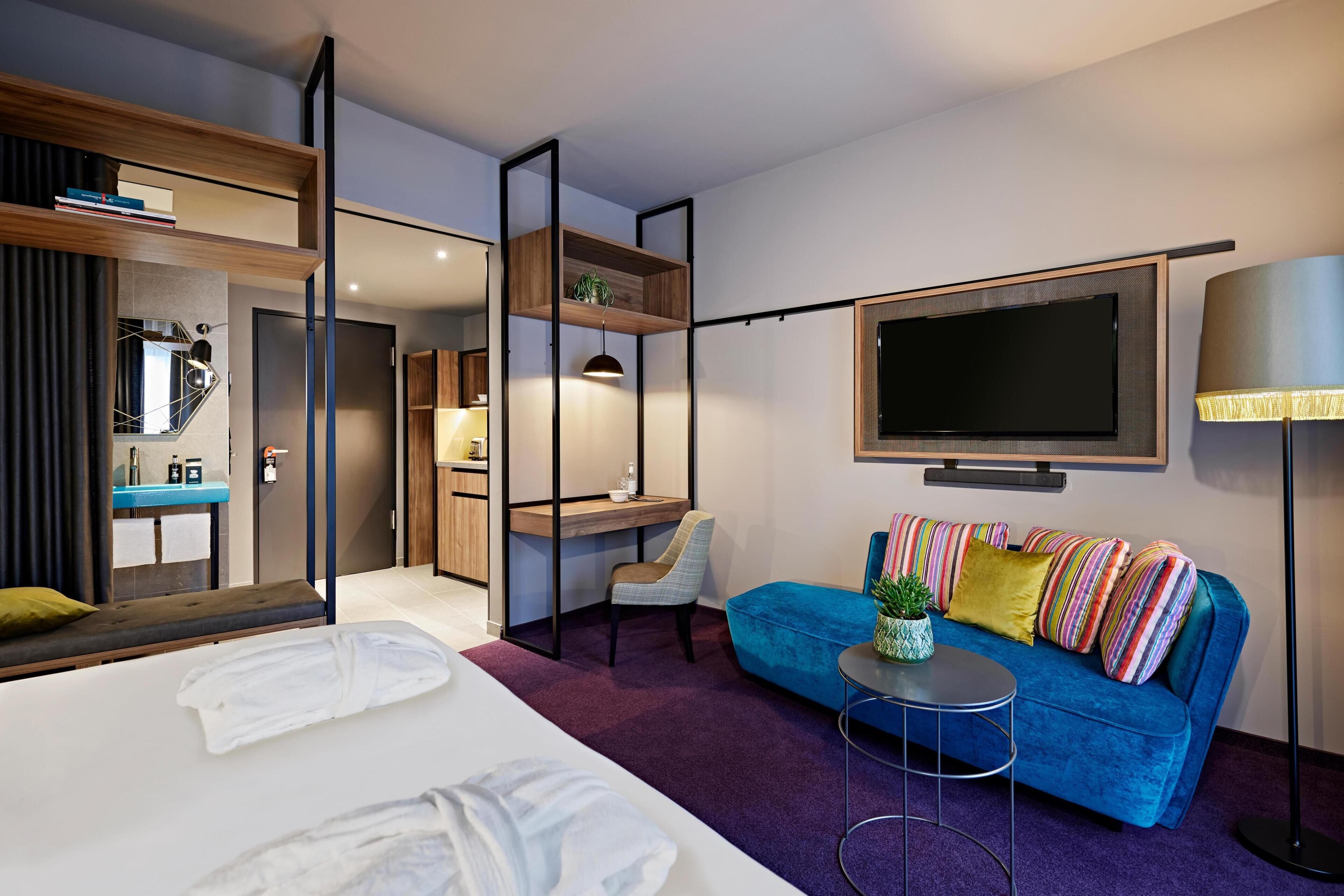 A modern hotel room with a bed, a blue sofa, a TV, and a desk with a chair.
