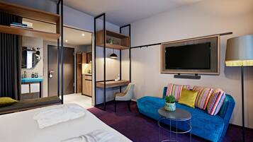 A modern hotel room with a bed, a blue sofa, a TV, and a desk with a chair.