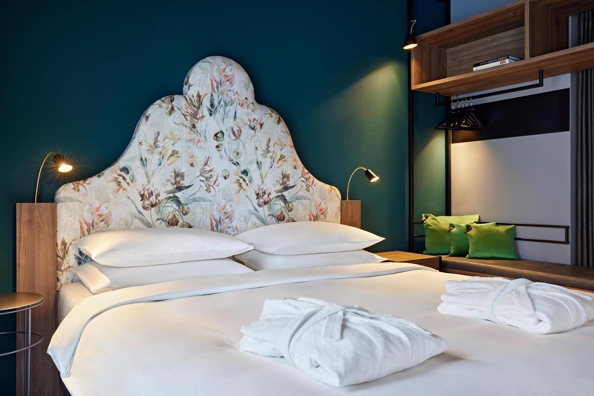 A neatly made bed with white linens, a floral headboard, and a pair of folded towels on the bed.