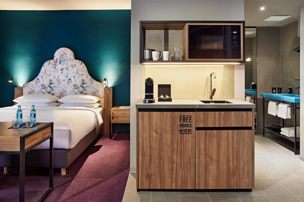 A hotel room with a bed, a wooden cabinet with a sign indicating free drinks, a nightstand, a bedside table with bottles, and a blue countertop with a sink.