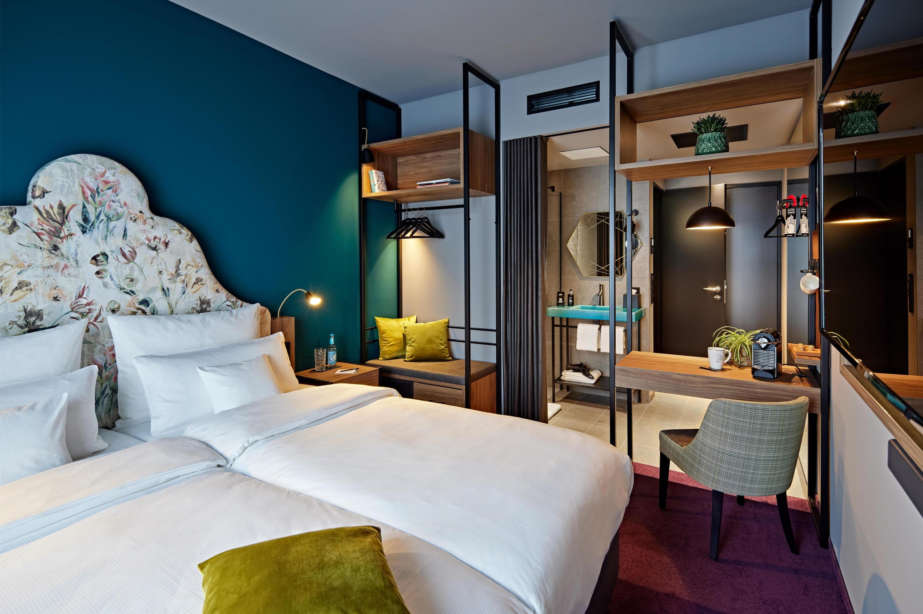 A hotel room with a bed, a desk, a chair, a mirror, and shelves with plants.