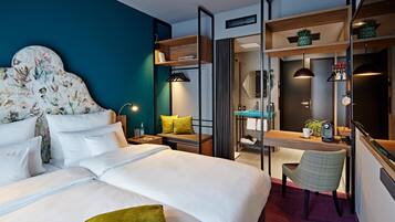 A hotel room with a bed, a desk, a chair, a mirror, and shelves with plants.