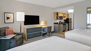 Ein Hotelzimmer mit zwei Betten, einem Schreibtisch mit Stuhl, einem Fernseher, einer Küchenzeile und einem gerahmten Bild an der Wand.