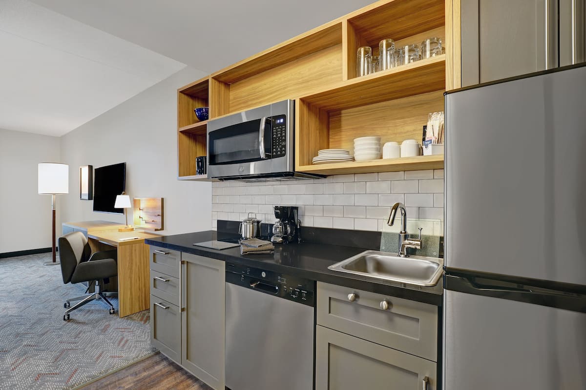 Una cocina moderna con microondas, cafetera y lavabo. Hay un estante de madera sobre el microondas y un lavavajillas debajo.