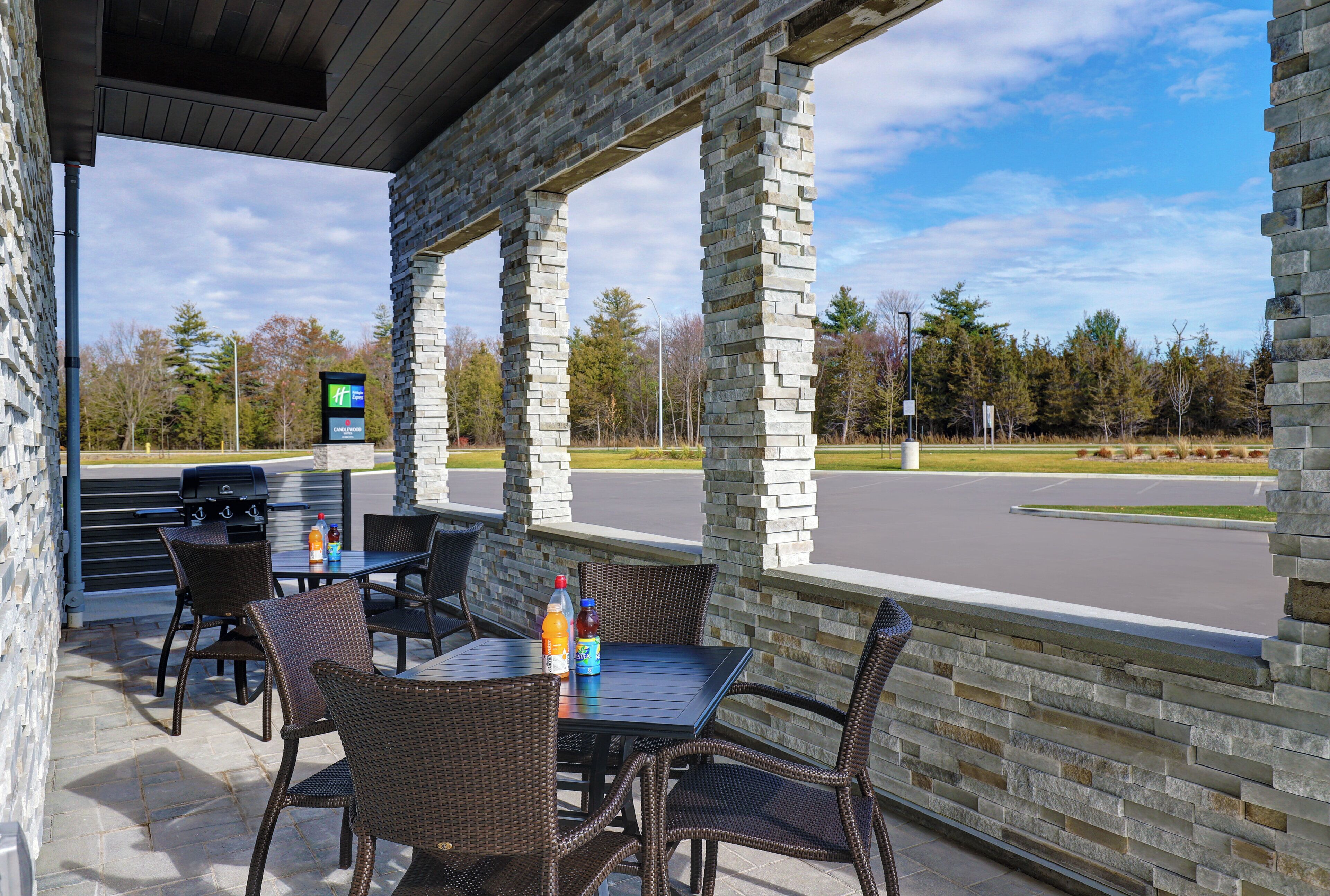 Un espace extérieur aménagé en terrasse, avec des piliers en pierre, des meubles en osier et une vue sur un parking et des arbres.