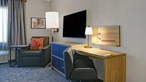 Ein Hotelzimmer mit Schreibtisch, zwei Stühlen, einem Flachbildfernseher, einer Lampe, einem Bett und einem Fenster mit Vorhängen.