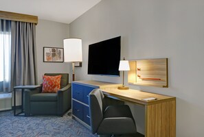 Habitación de hotel con escritorio, dos sillas, televisión de pantalla plana, lámpara, cama y ventana con cortinas.