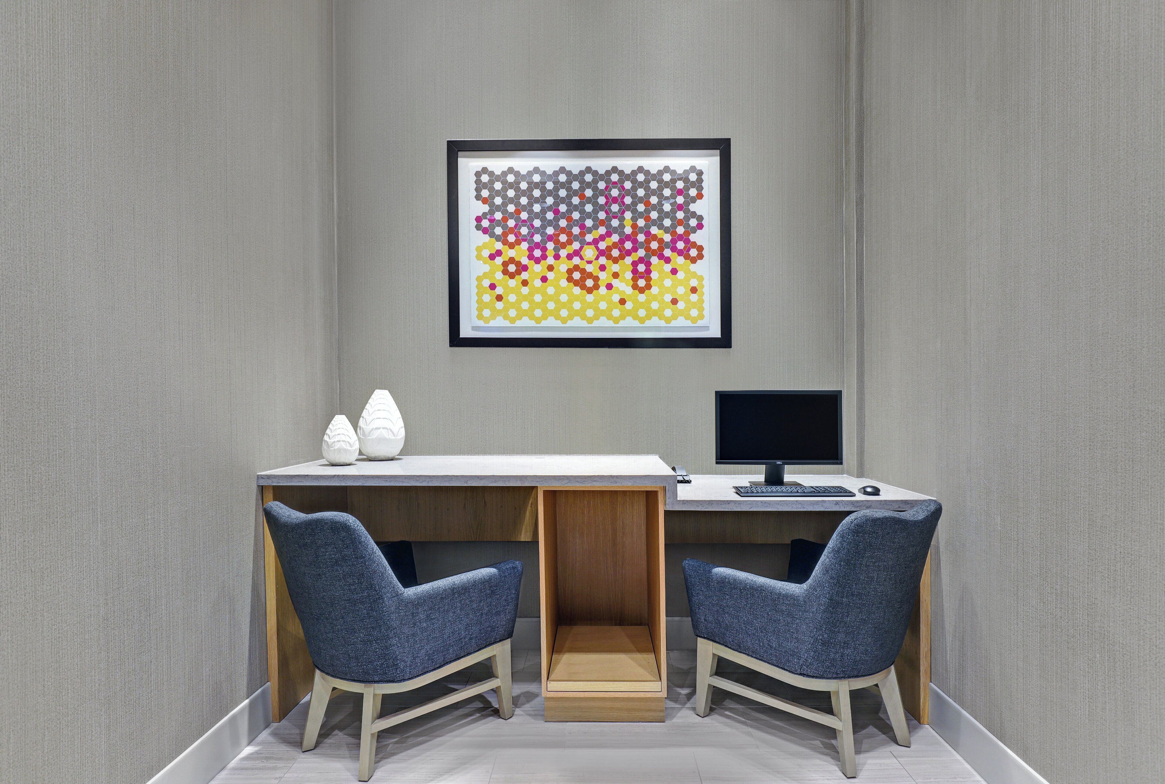 A room with a desk, two chairs, a computer, and a framed artwork on the wall.