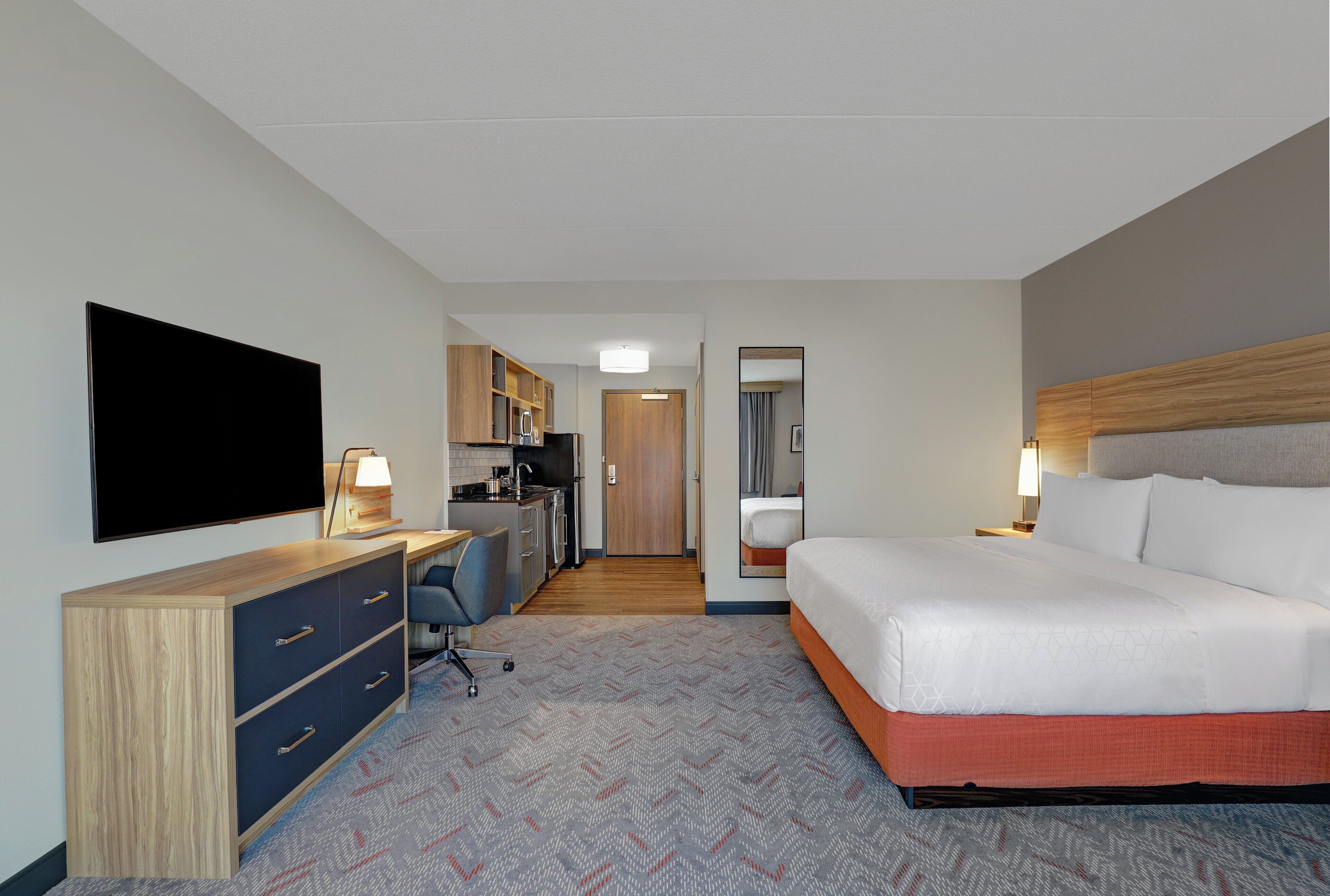 A hotel room with a bed, desk, chair, television, and kitchenette.