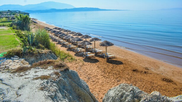 A beach with rows of sun loungers and umbrellas, a clear sky, and distant mountains. - Xi Village (Kefalonia)