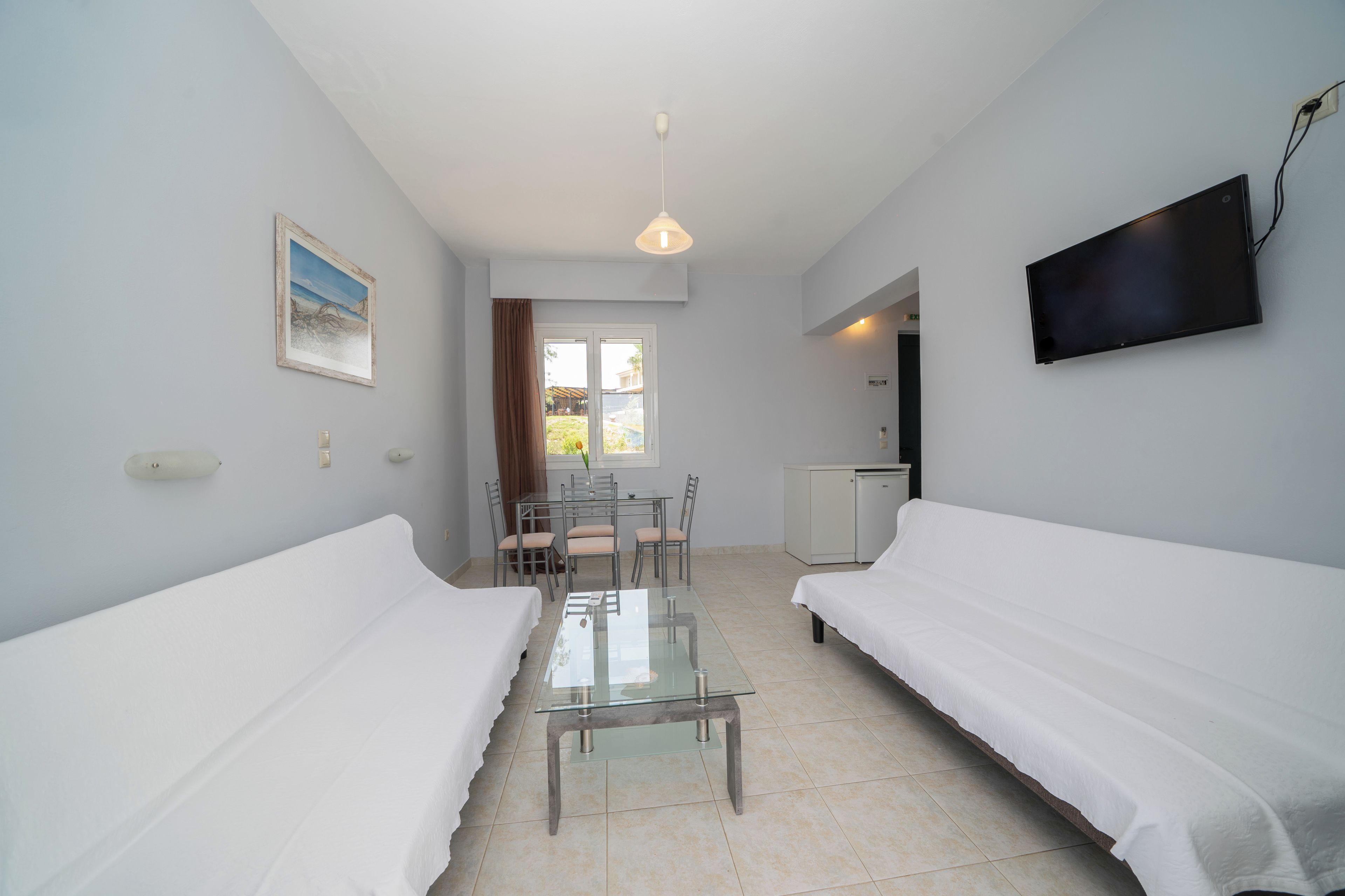 A room with two white beds, a glass-top table, a TV mounted on the wall, and a window with curtains.