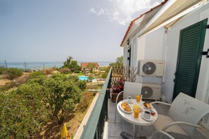 A balcony with a table set for two, overlooking a pool and beach. - Xi Village (Kefalonia)