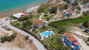 An aerial view of a coastal property with a swimming pool, multiple buildings, and a beach area. - Xi Village (Kefalonia)