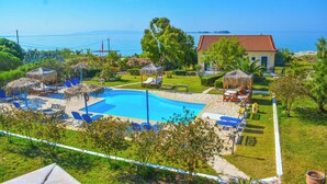 A pool area with lounge chairs and umbrellas, surrounded by greenery and a building with a red roof. - Xi Village (Kefalonia)
