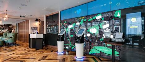 A modern hotel lobby with a herringbone wood floor, a bar area with green upholstered chairs, and a large digital map display.