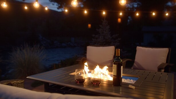 Une table avec une bouteille et des verres, un feu allumé et des guirlandes lumineuses, le tout dans un décor extérieur nocturne.