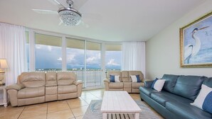 A spacious living room with a beige sectional sofa, a dark green sofa, and a white coffee table. There is a large window offering a view of the city skyline.