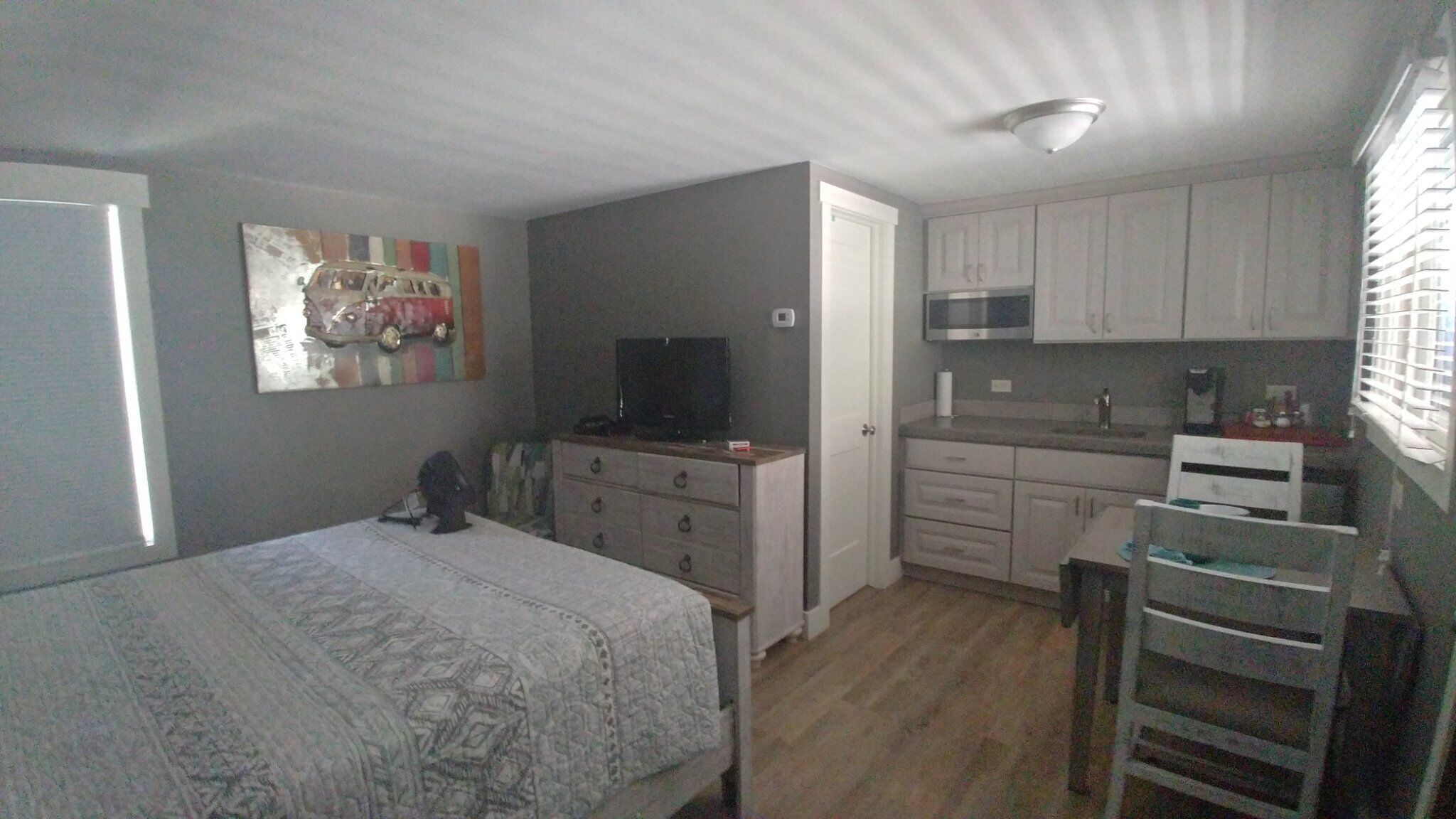 A bedroom with a bed, a dresser, and a television. There is a kitchenette with a microwave and cabinets.