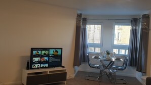 A living room with a flat-screen TV on a stand, a dining table with chairs, a window with curtains, and a small white cabinet. - Beach Apartment - Kirkcaldy (Kirkcaldy)