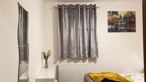 A bedroom with a bed, a radiator, a window with grey curtains, a mirror, and a painting on the wall. - Beach Apartment - Kirkcaldy (Kirkcaldy)