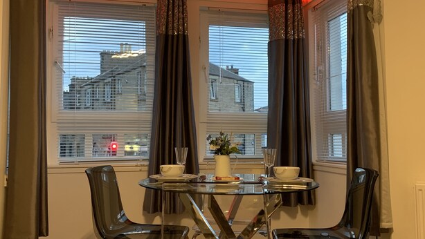 A corner room with two windows, a table with chairs, and a vase with flowers. - Beach Apartment - Kirkcaldy (Kirkcaldy)