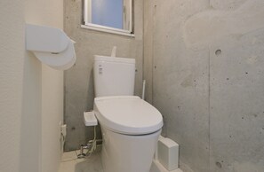 A small, compact bathroom with a white toilet, a visible water pipe, and a window above the toilet. - Landmark Namba C (Osaka)