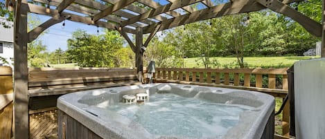 Un bain à remous sous une pergola en bois, avec une terrasse en bois et une pelouse verdoyante en arrière-plan.