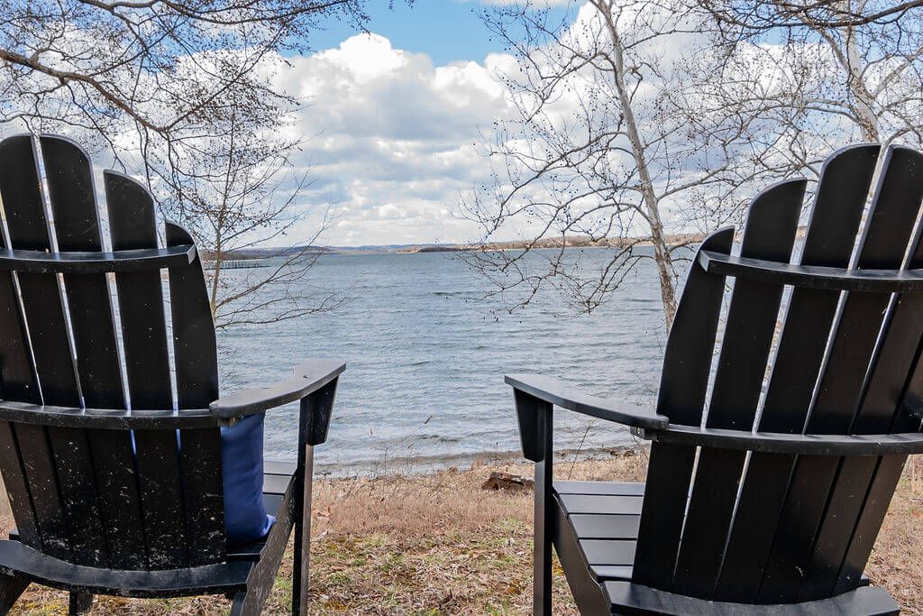 Two Adirondack chairs facing a body of water with a distant shoreline and trees in the background.