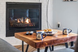A living room with a fireplace, a wooden table with mugs and a plate of food, and a TV mounted on a white brick wall.