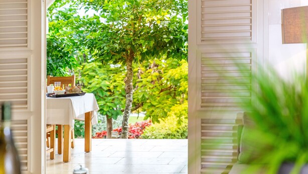 A room with a view of a lush garden, a dining table set with breakfast, and a sofa. - Maison Cerisier accès mer (Le François)