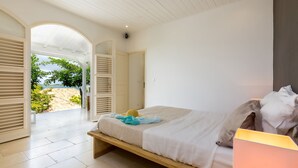 A bedroom with a bed, a bedside lamp, and an open door leading to a balcony with a view of the sea. - Maison Cerisier accès mer (Le François)