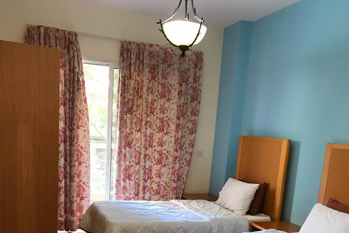 A hotel room with two beds, a wooden bench, and a large window with floral curtains.