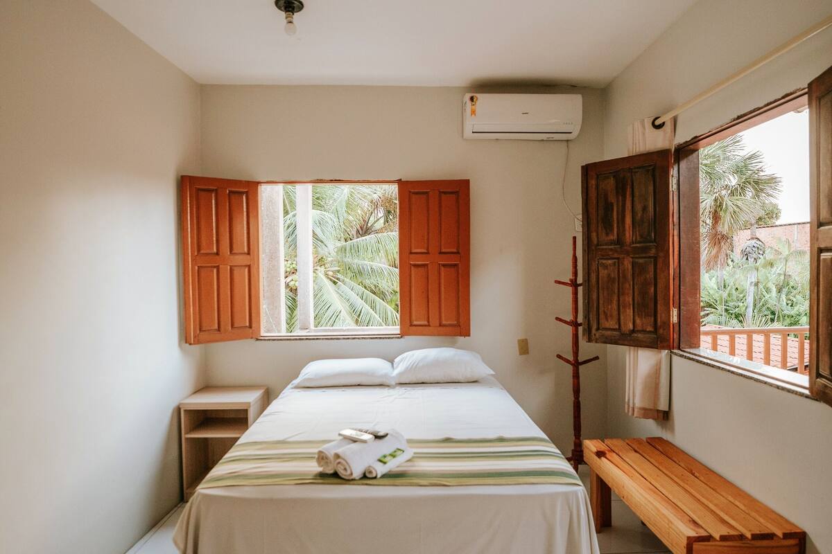 Un dormitorio con cama, un banco de madera, una ventana con contraventanas abiertas y vistas a la vegetación exterior.