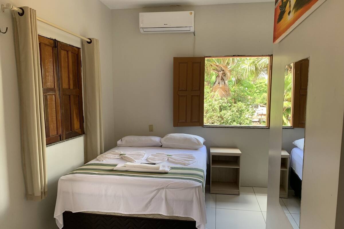 Un dormitorio con cama, ventana con vista a la vegetación, mesita de noche pequeña y equipo de aire acondicionado.