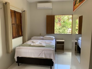 A bedroom with a bed, a window with a view of greenery, a small bedside table, and an air conditioning unit.
