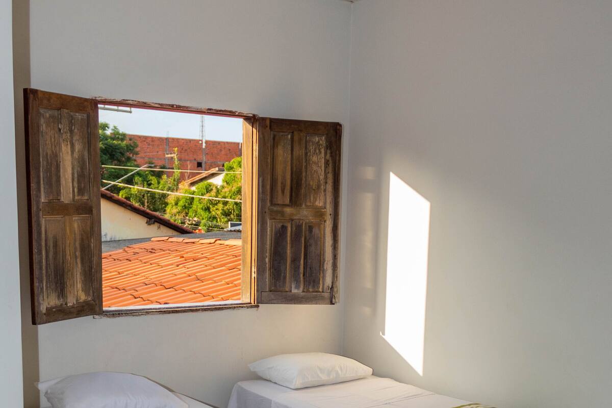 Una habitación con dos camas individuales, una ventana con contraventanas de madera y una lámpara en el techo.