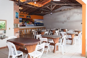 A restaurant with wooden tables and white plastic chairs, a brick wall, and a tiled floor.