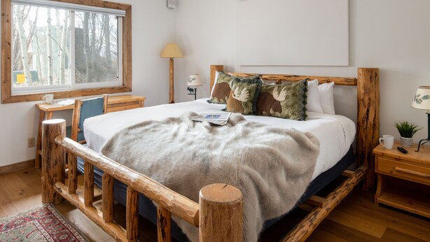 A bedroom with a wooden bed frame, a large window with a view of trees, a bedside table with a lamp, and a small plant on the nightstand. - Outpost: Pitchfork 434 - 1 bedroom near Snow King! (Jackson)