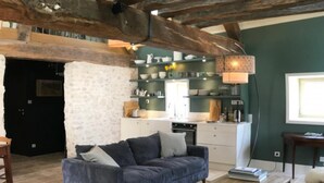 A rustic interior with exposed wooden beams, a stone wall, a sofa, and a kitchen area with shelves and appliances. - Colivault (Cande-sur-Beuvron)