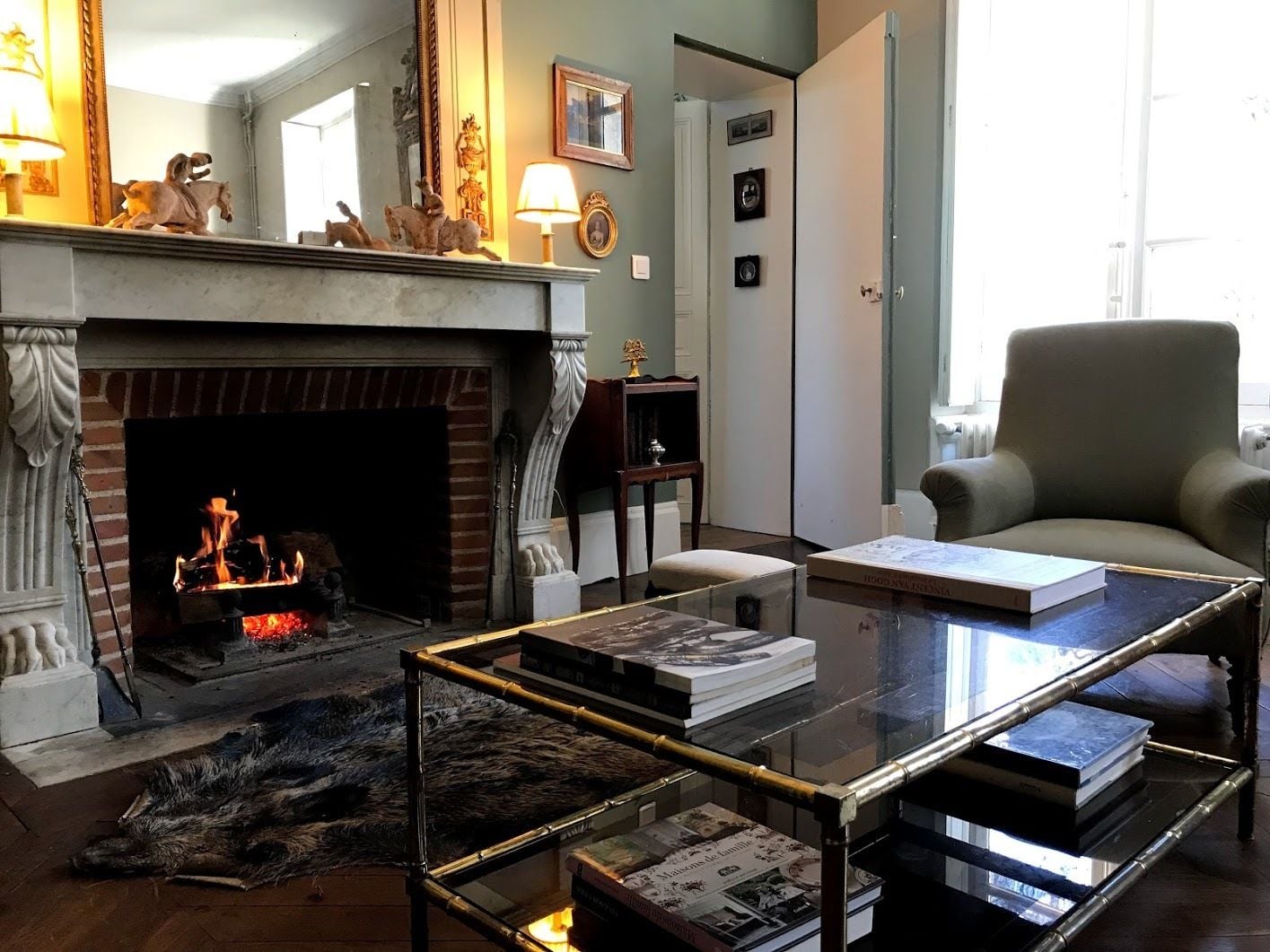 A living room with a fireplace, a glass coffee table with books, a comfortable armchair, and a lamp.