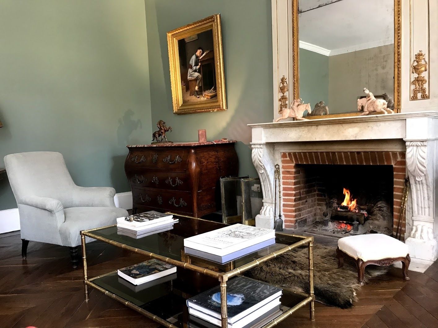 Una sala de estar con chimenea, un sillón blanco, un aparador de madera y una mesa de centro de vidrio con libros.