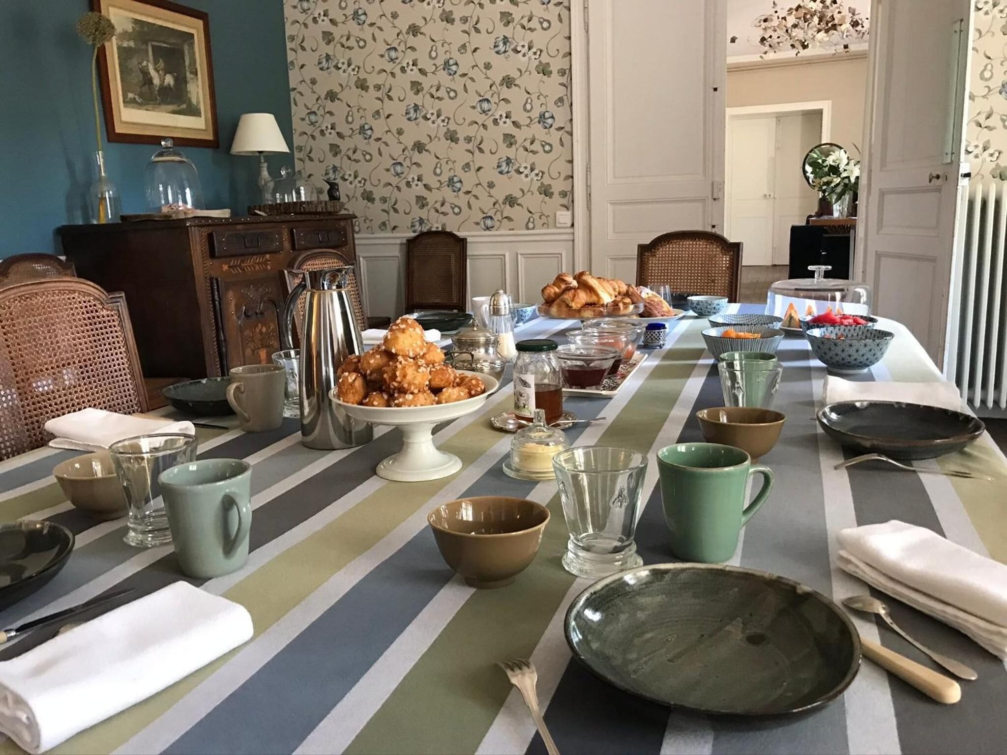 A dining table set with various dishes, glasses, and utensils, surrounded by wicker chairs and a patterned wallpaper.